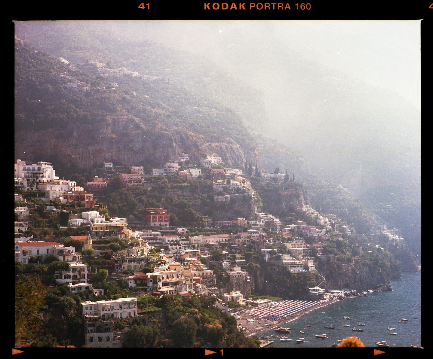 A town with many buildings is built into a steep hillside overlooking a beach and the sea.