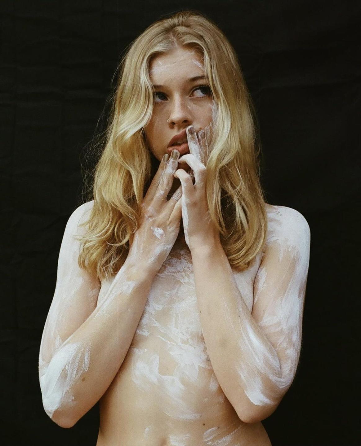 A woman with long blonde hair and white paint on her body looks to the side against a dark background.