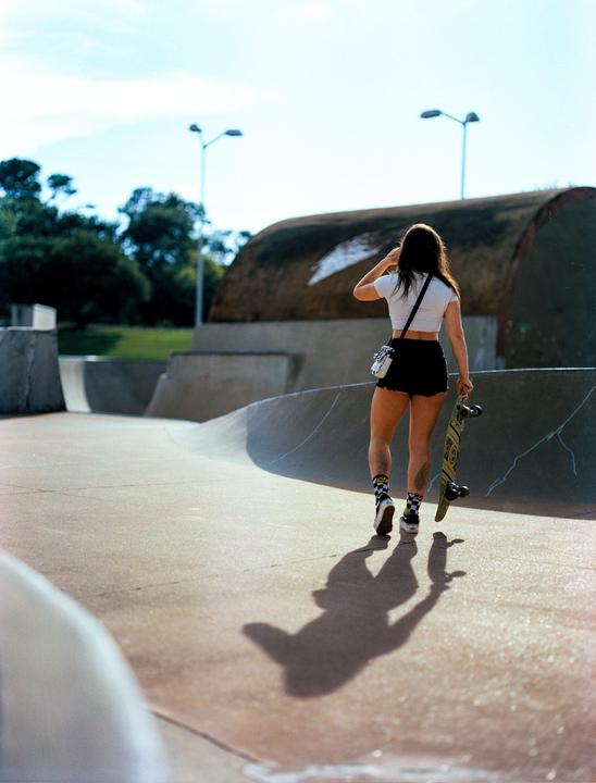 A woman walks away from the viewer, carrying a skateboard in a skatepark.