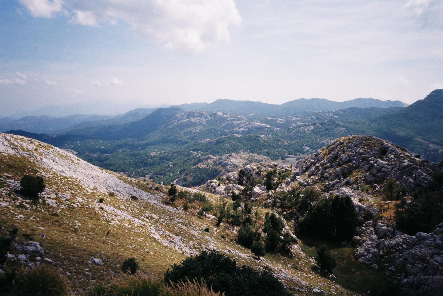 A vast mountainous landscape with rocky slopes and scattered trees stretches under a cloudy sky.