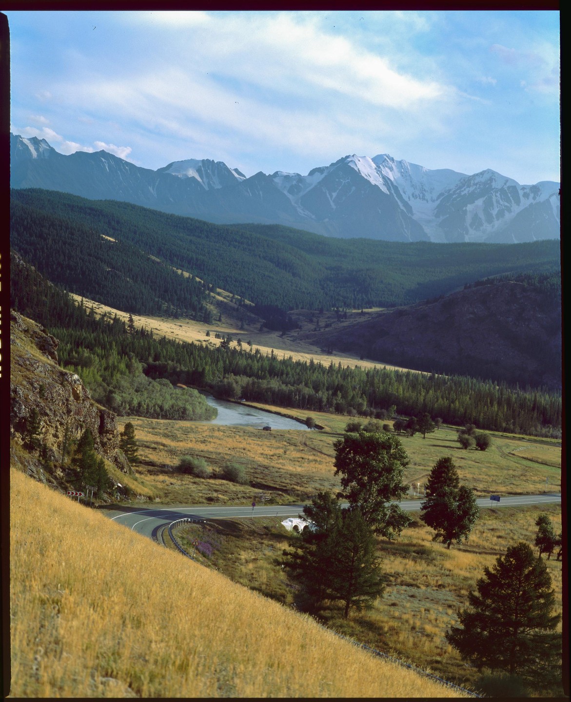 A winding road follows a river through a valley with forested hills and snow-capped mountains under a blue sky.
