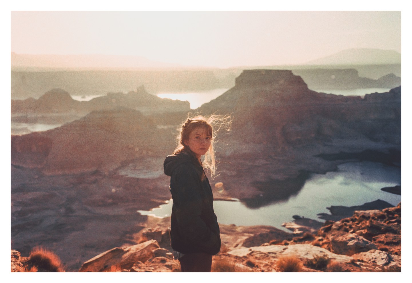 A woman stands on a rocky outcrop overlooking a vast desert landscape with a lake and mesas at sunset.