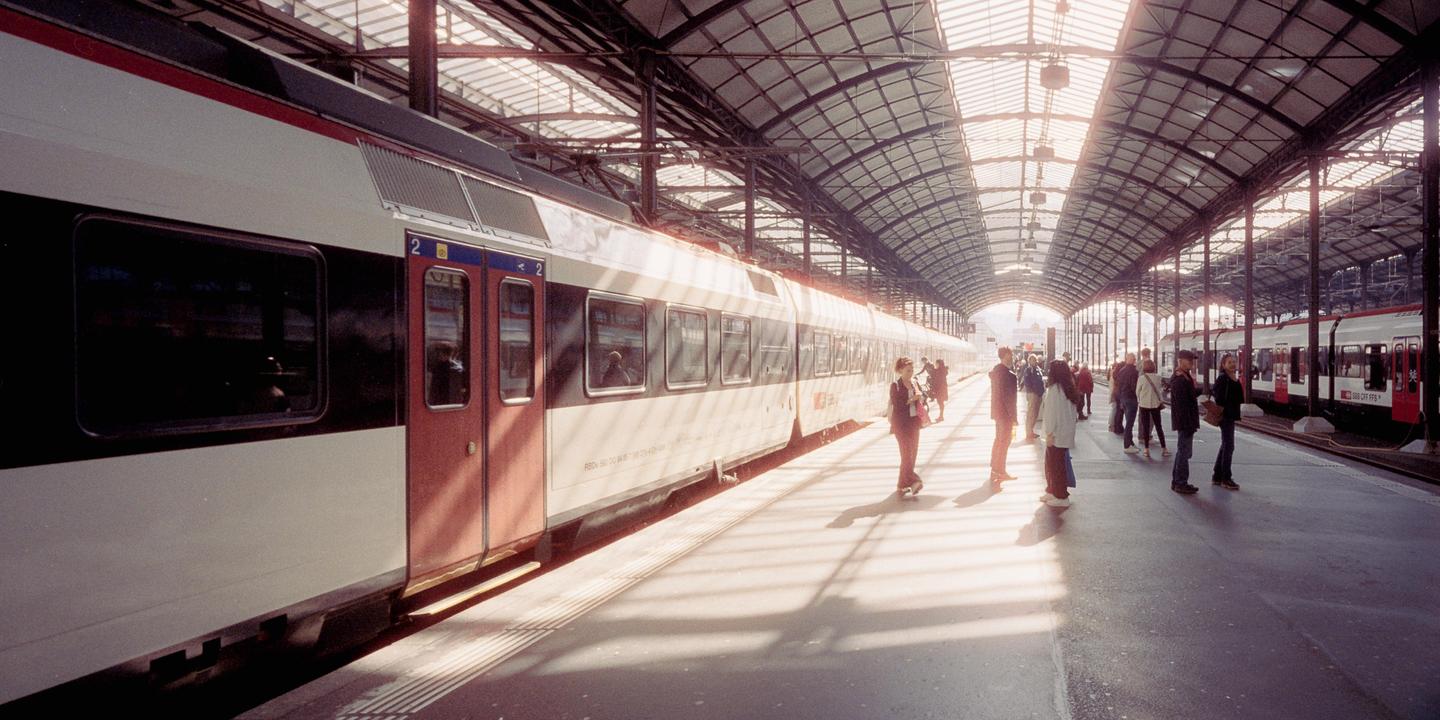People wait on a train platform next to a parked train under a glass and steel roof.