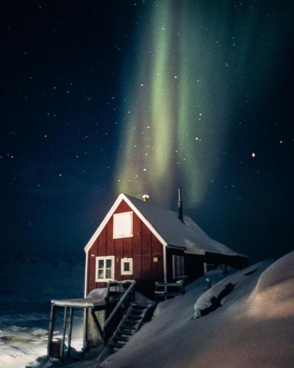 A red house covered in snow is illuminated by the aurora borealis under a starry night sky.