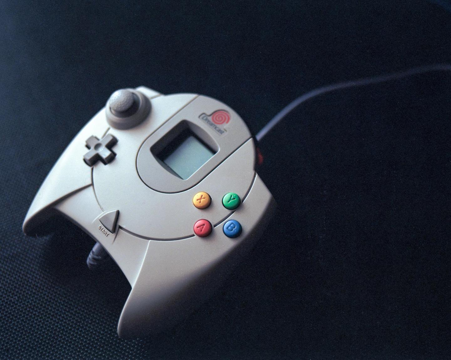 A Dreamcast controller with colorful buttons and a small screen is angled on a dark surface.