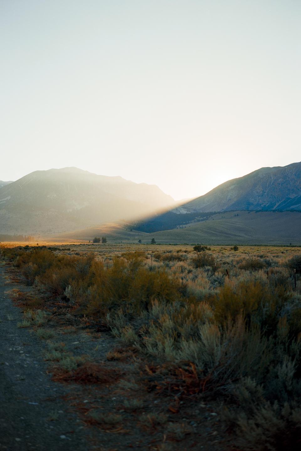 Sunlight streams across a vast, arid landscape with mountains in the background and scrub brush in the foreground.