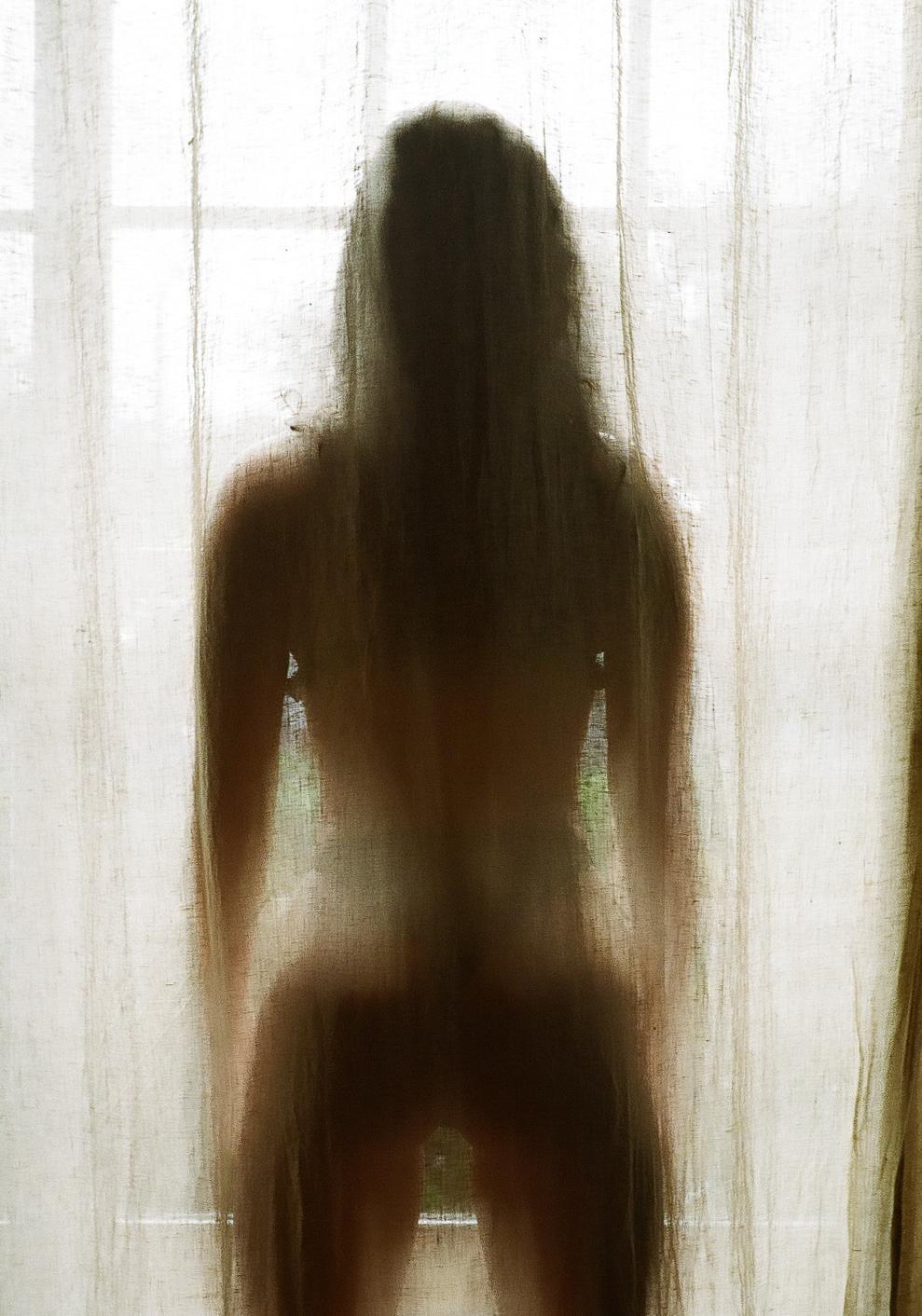 A woman stands behind a sheer curtain, her silhouette visible against the bright window.