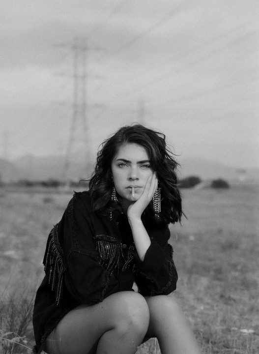 A woman with a cigarette in her mouth sits in a field with a power line in the background.