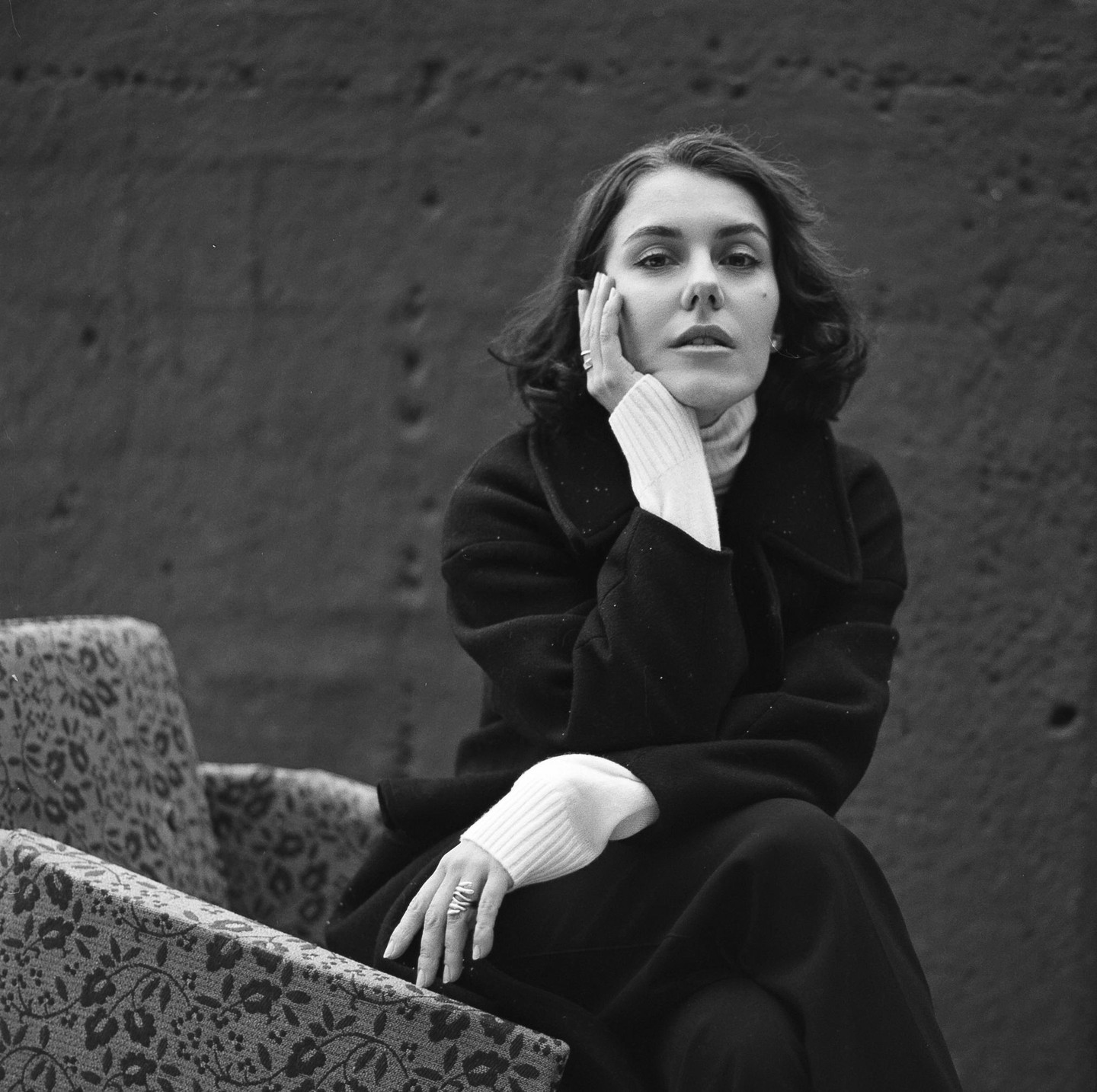 A woman in a dark coat and light sweater sits with her hand on her face next to a patterned chair.