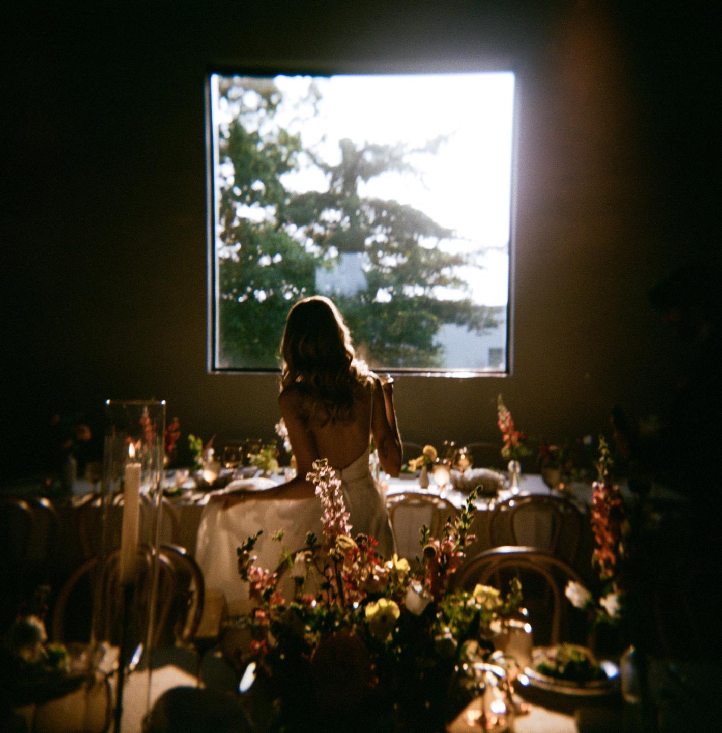 A woman in a white dress stands at a table with floral arrangements, looking out a window.