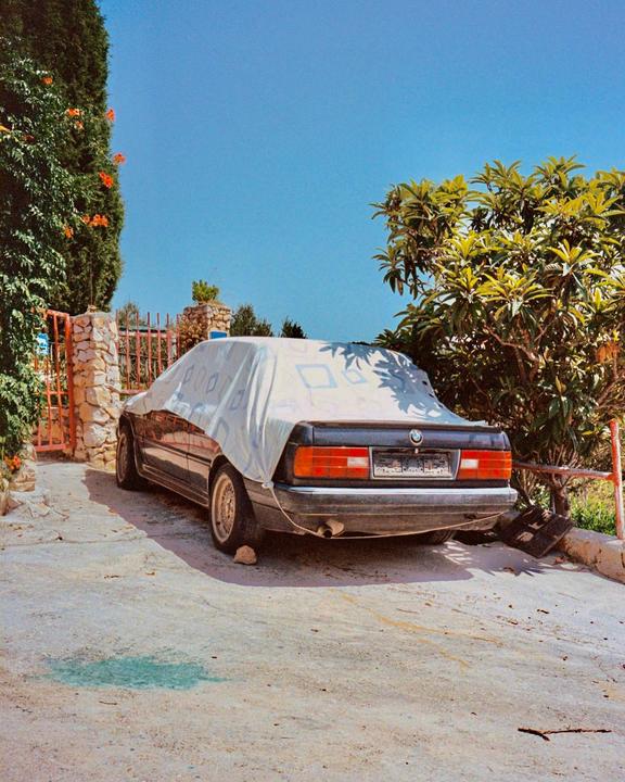 A black car covered with a patterned sheet is parked on a concrete driveway next to a stone wall and green foliage under a clear blue sky.