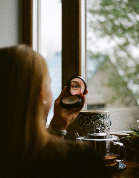 A woman holds a compact mirror to her eye, reflecting it, with a window and teapot in the background.