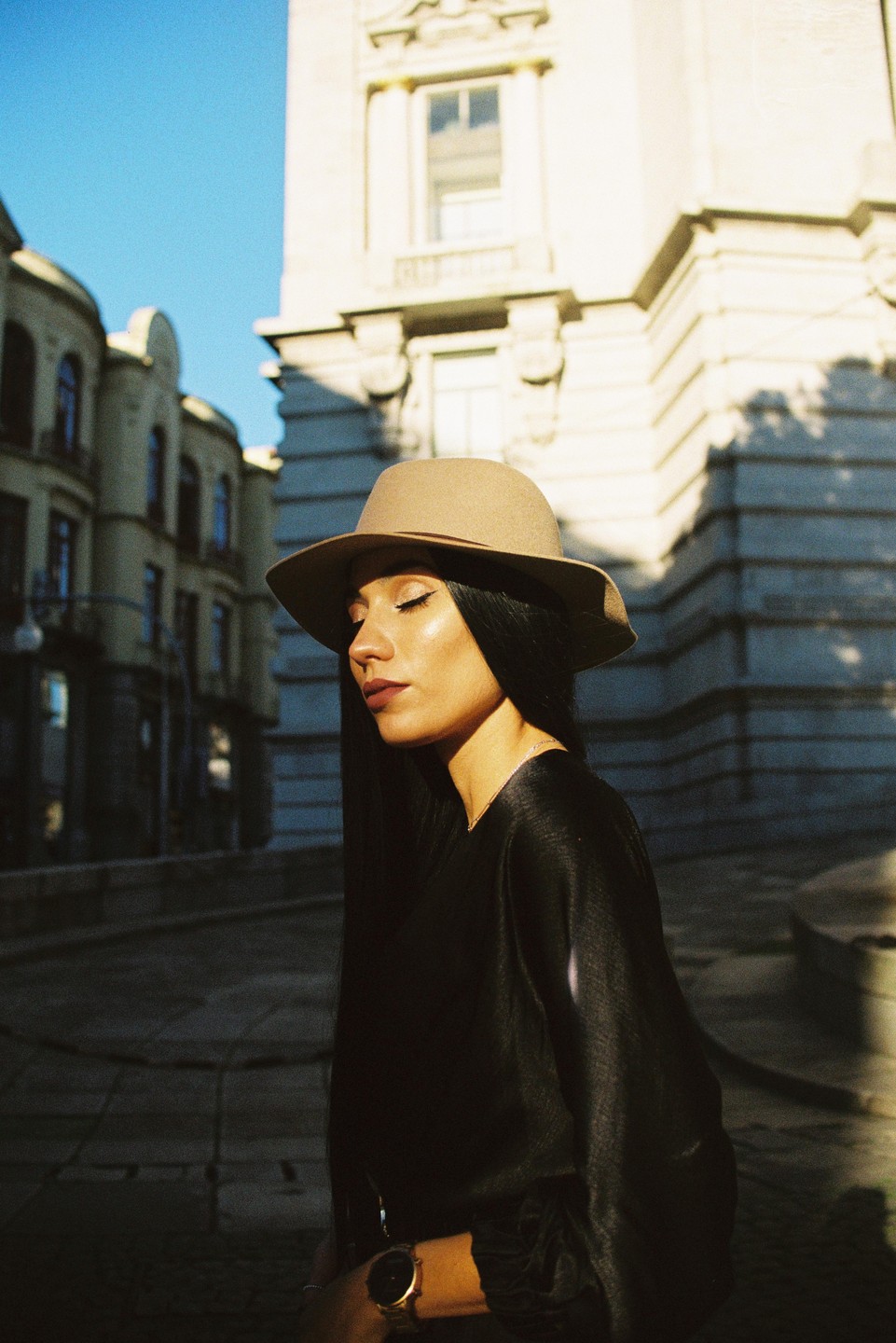 A woman wearing a hat and a black top stands on a street with buildings in the background, illuminated by sunlight.