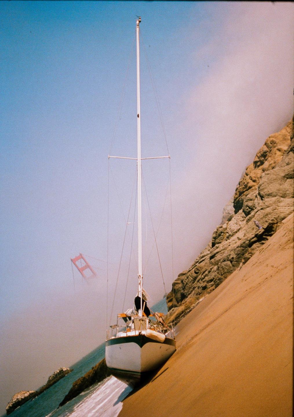 A sailboat is beached on a sandy shore next to a rocky cliff, with the Golden Gate Bridge visible through fog in the distance.