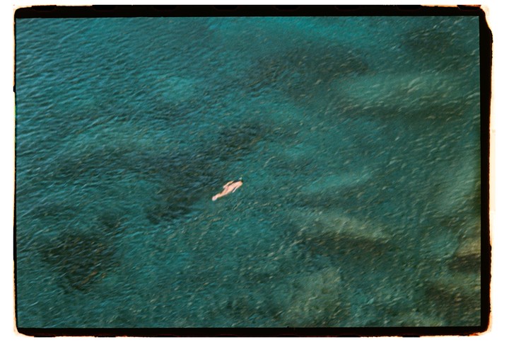 A person swims in clear blue water, seen from above.