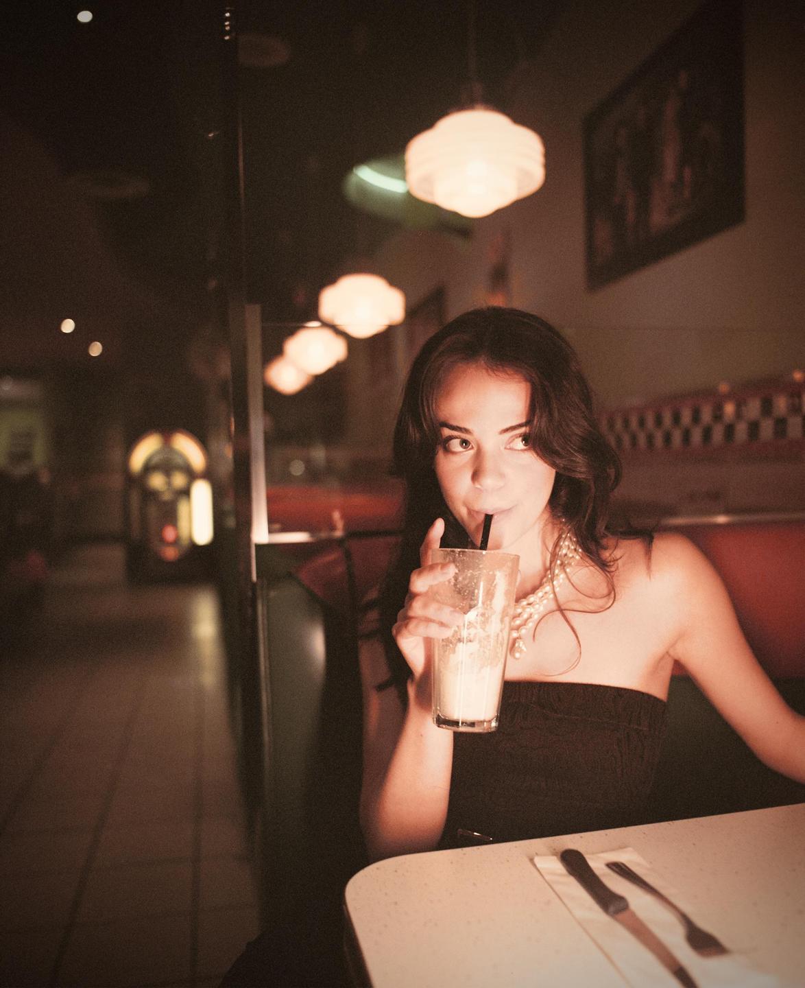A woman sips a milkshake in a diner booth.