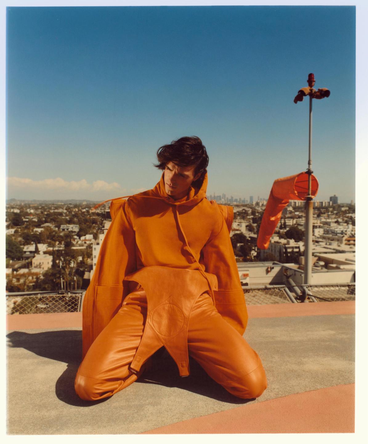 A man in an orange outfit kneels on a rooftop with a cityscape in the background.