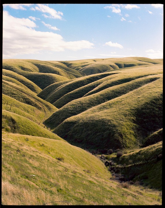 Rolling green hills are illuminated by sunlight under a blue sky with scattered clouds.
