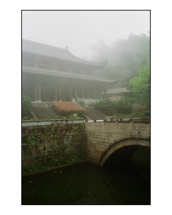A traditional temple with a curved roof stands behind a stone bridge over a canal, all shrouded in mist.