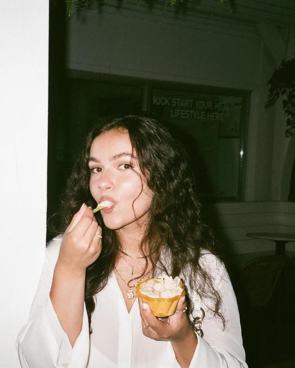A woman with long, curly hair eats gelato from a yellow cup with a green spoon.