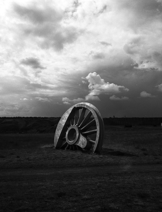 A large metal wheel sculpture is partially buried in a grassy field under a cloudy sky.