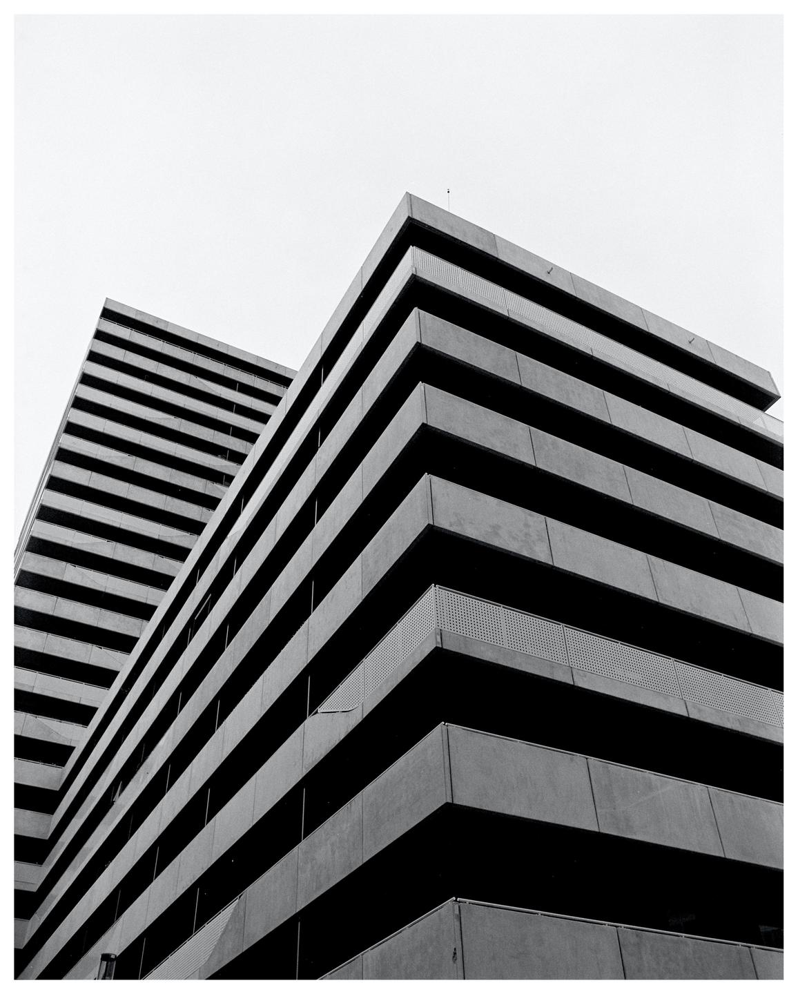 Two brutalist buildings with horizontal lines are seen from a low angle against a bright sky.
