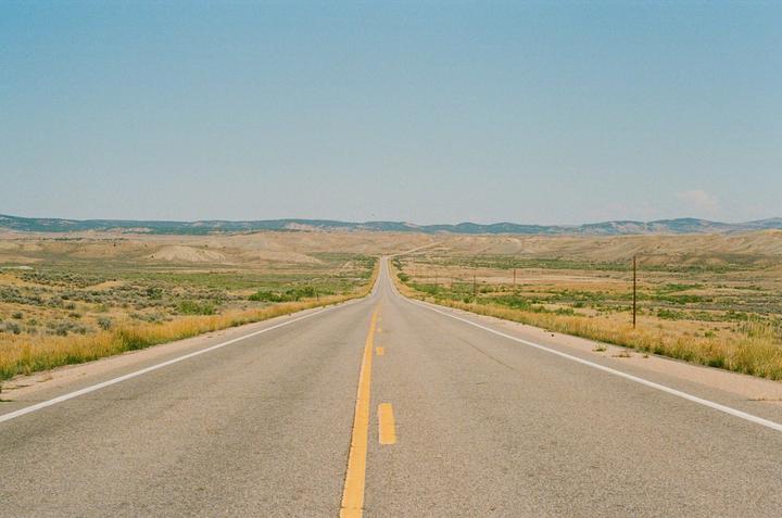 A long, straight road with a yellow dividing line extends into the distance through a dry, grassy landscape under a clear sky.