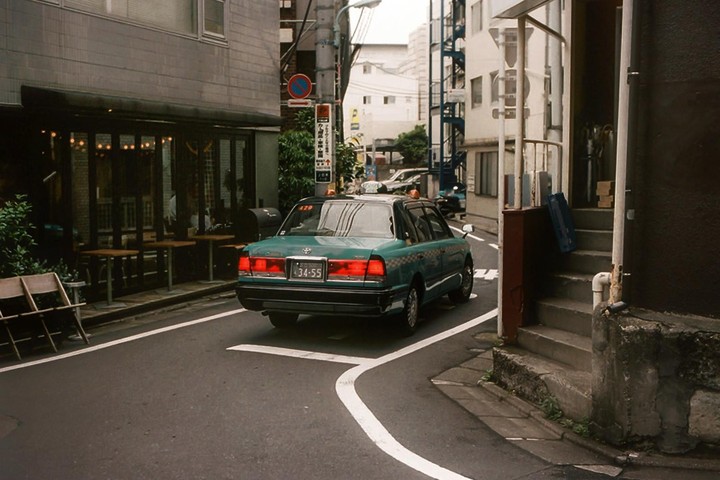 A green taxi drives down a narrow street past a cafe with outdoor seating.
