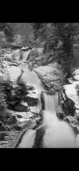 A long exposure captures a multi-tiered waterfall cascading over snow-covered rocks, with a wooden bridge visible in the background.