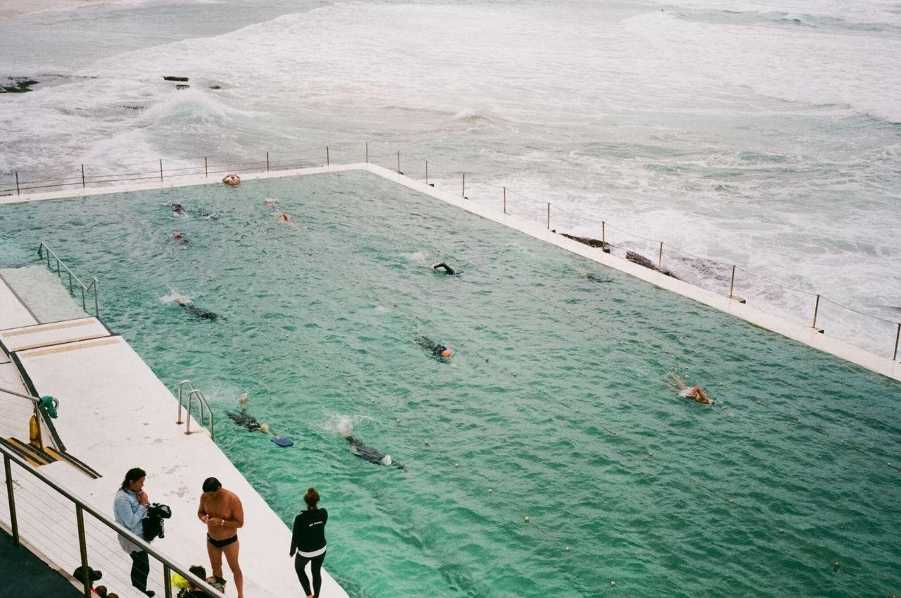 A group of people swim in a large pool next to the ocean while others stand on the pool deck.