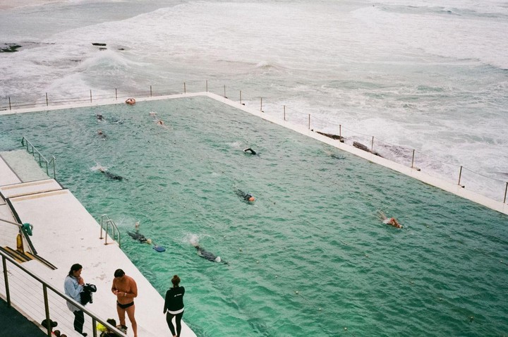 A group of people swim in a large pool next to the ocean while others stand on the pool deck.