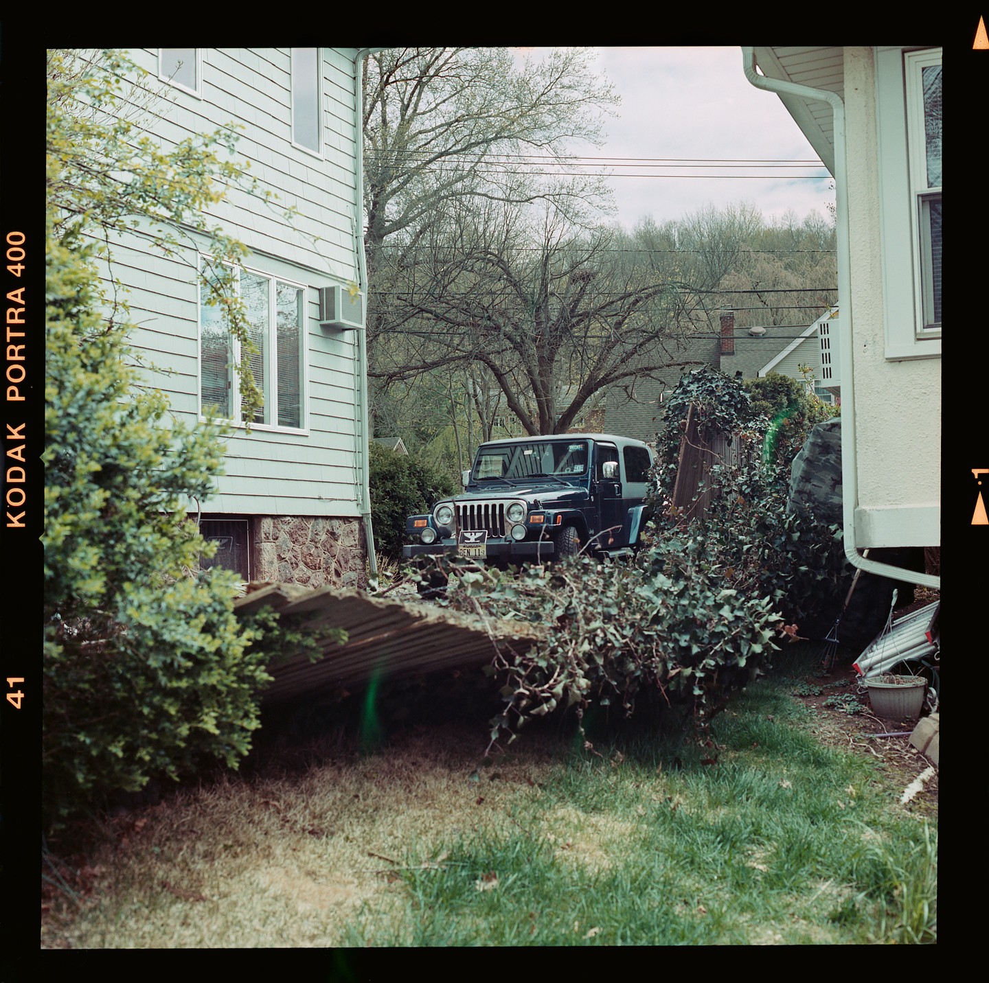 A dark blue jeep is parked between two houses, surrounded by overgrown bushes and trees.