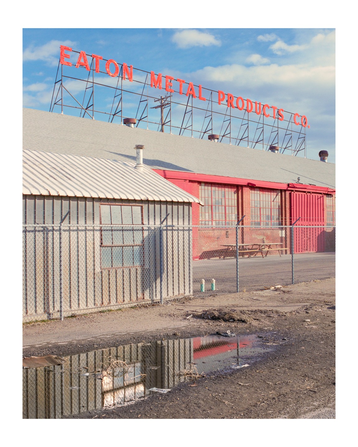 A red sign for Eaton Metal Products Co. is visible above a building with a puddle reflecting the building and sky in the foreground.
