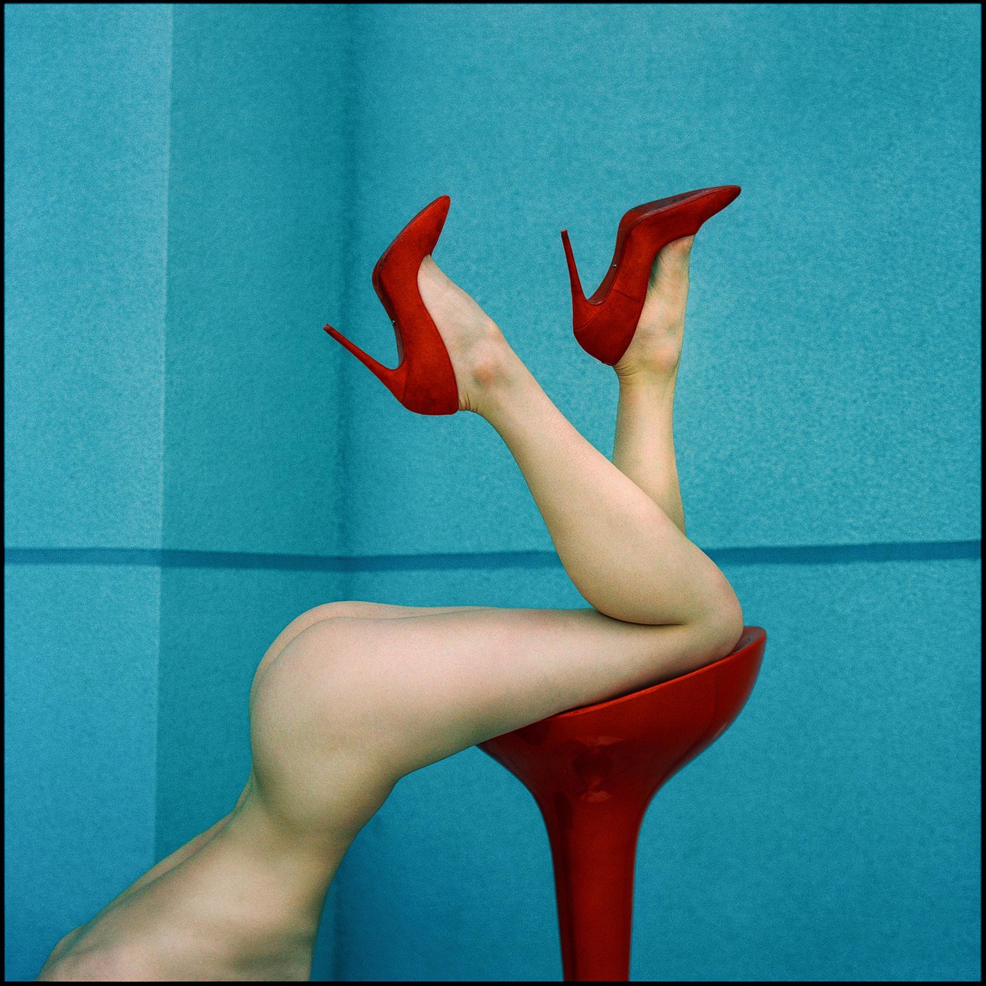A woman's legs and bare bottom are visible as she lies on a red stool, wearing red high heels against a blue background.