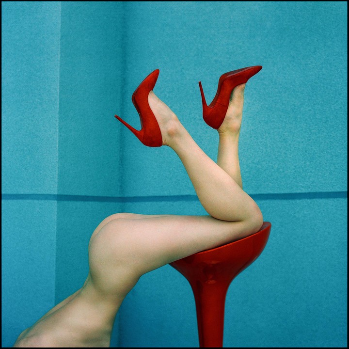 A woman's legs and bare bottom are visible as she lies on a red stool, wearing red high heels against a blue background.