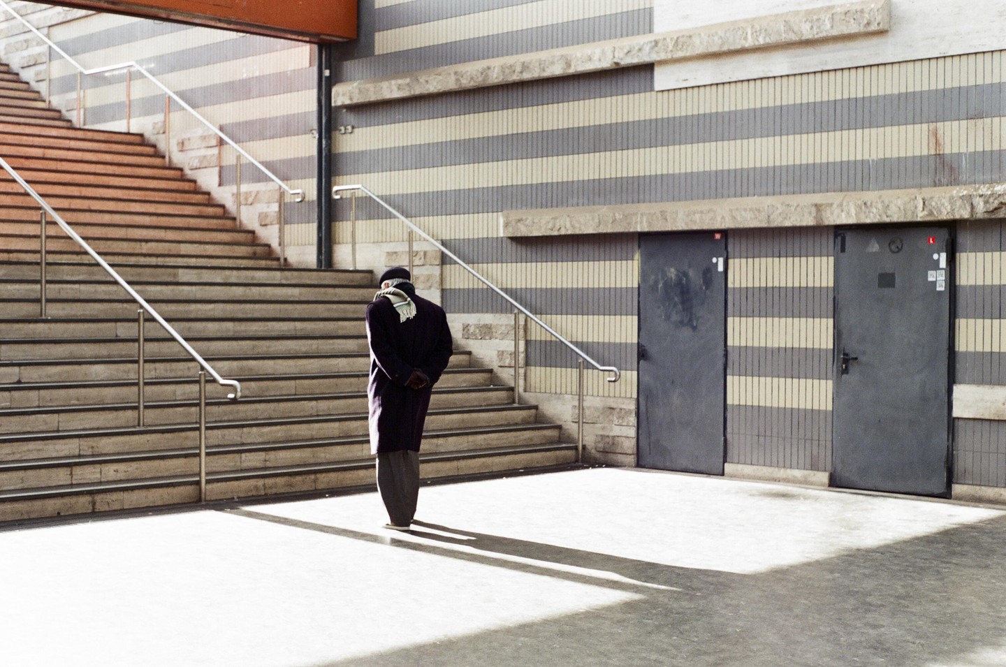 A man stands at the bottom of a wide staircase with two doors and a striped wall behind him.