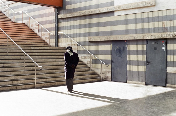 A man stands at the bottom of a wide staircase with two doors and a striped wall behind him.