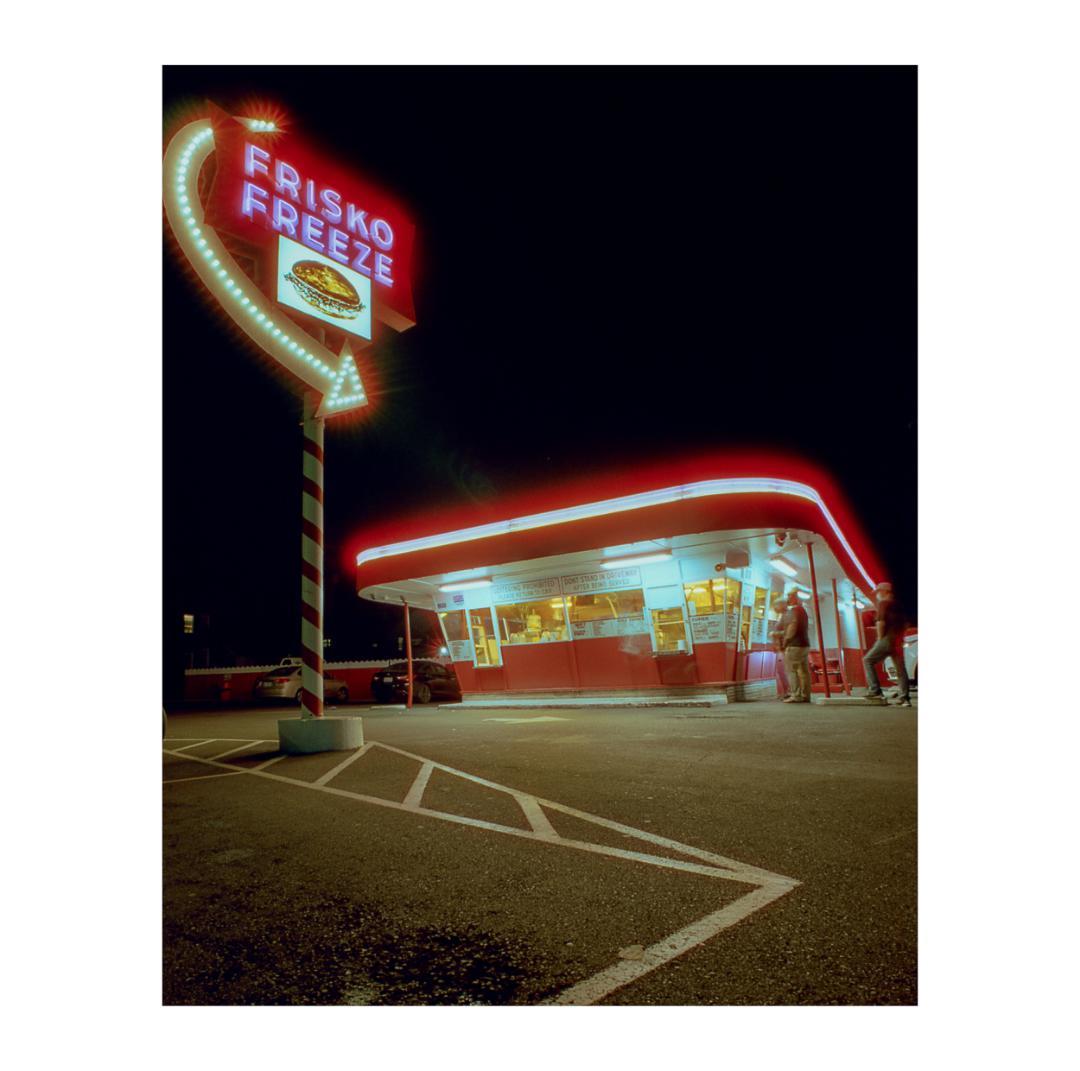 A neon sign for Frisko Freeze stands next to a brightly lit diner at night.