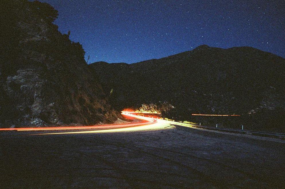 Light trails from vehicles illuminate a winding road at night, with mountains and a starry sky in the background.