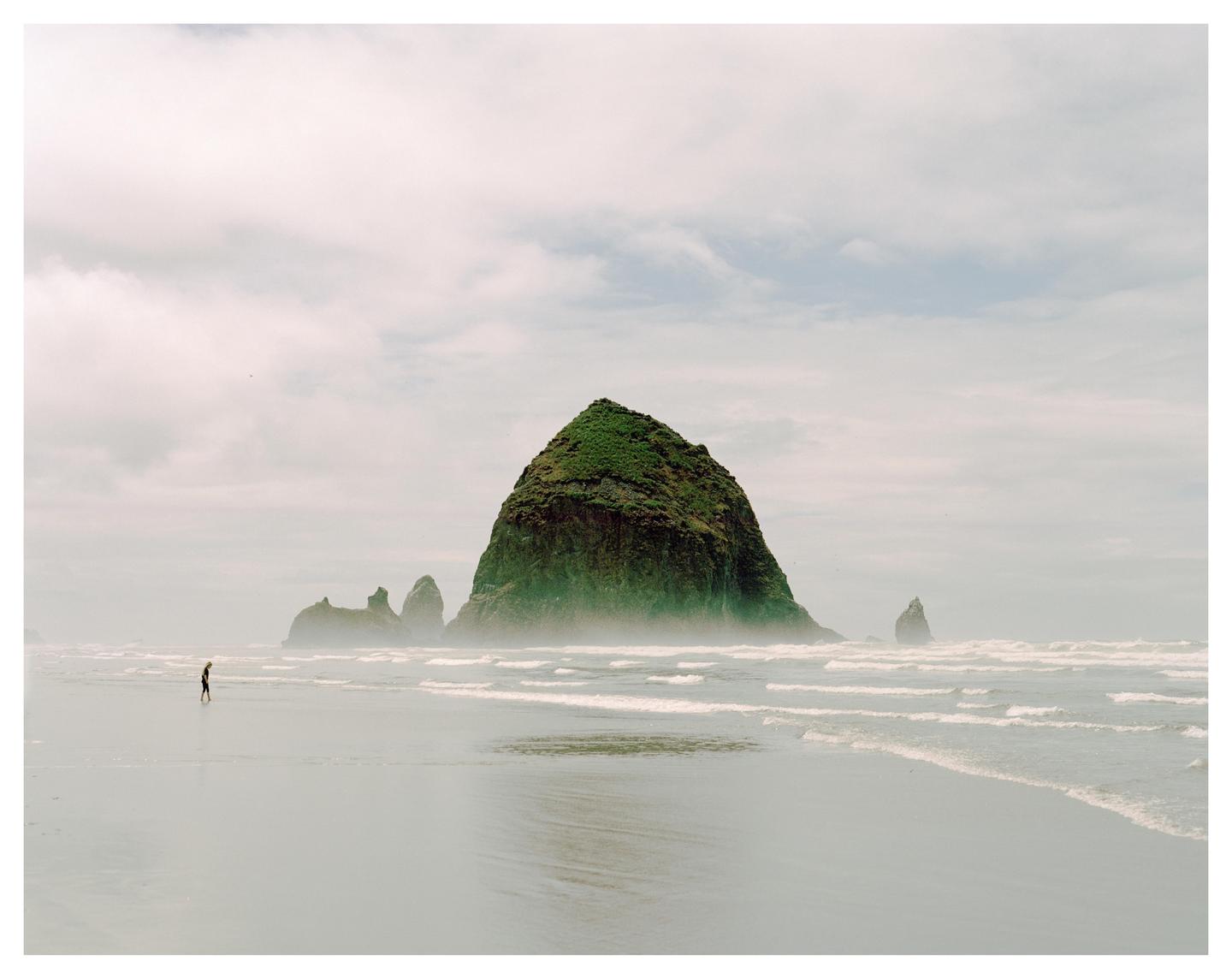 A person walks on a foggy beach with a large, green, rocky outcropping in the background.