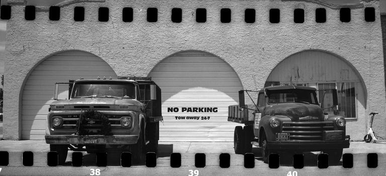 Two old trucks are parked in front of a building with three arched garage doors, the middle one displaying a "NO PARKING" sign.