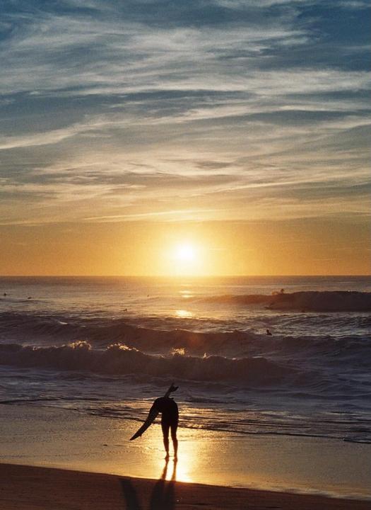 A surfer stands on the beach at sunset, holding a surfboard and looking out at the ocean waves.