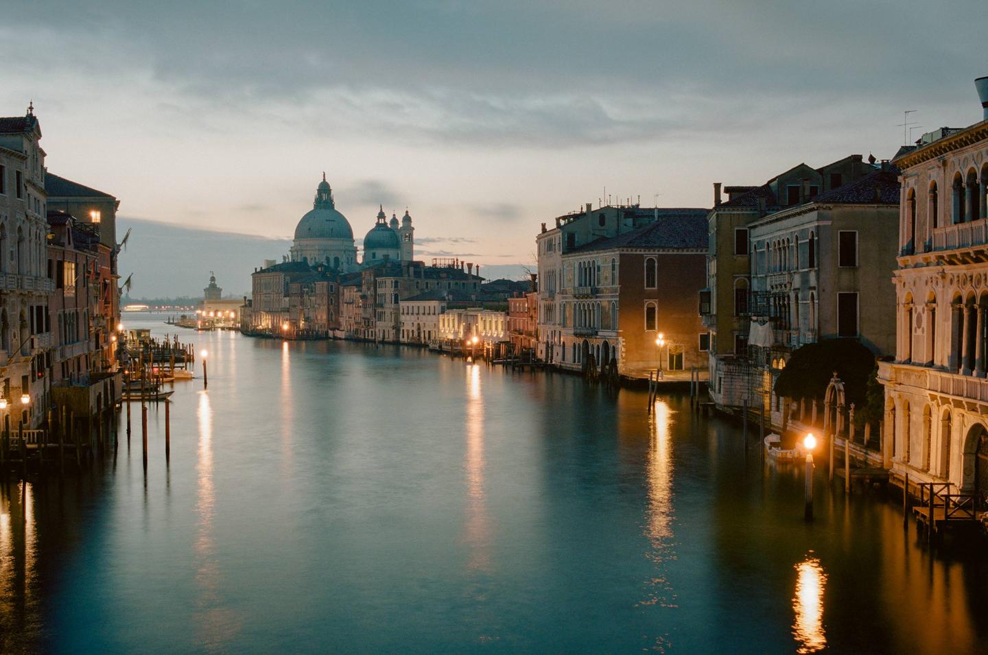 A canal with buildings on both sides and a large domed building in the distance is illuminated by streetlights reflecting on the water.