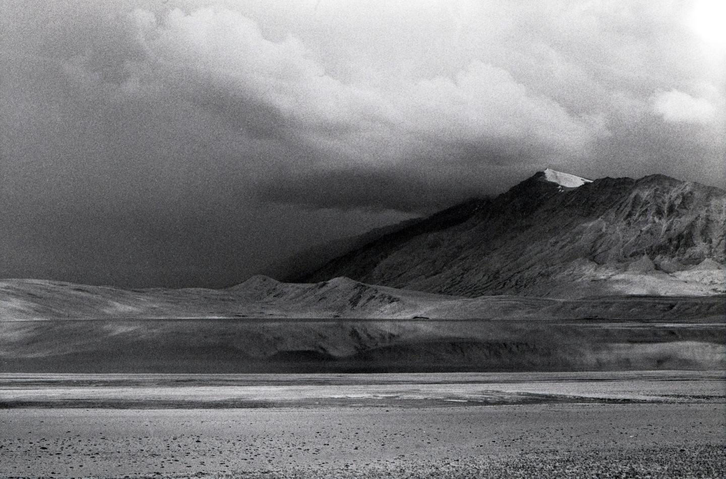 A mountain range with a snow-capped peak is reflected in a still body of water under a cloudy sky.
