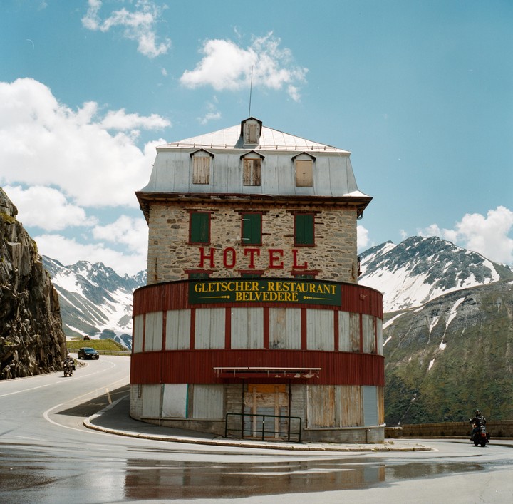 A stone hotel with a red base stands at a curve in a wet road, with snow-capped mountains in the background.