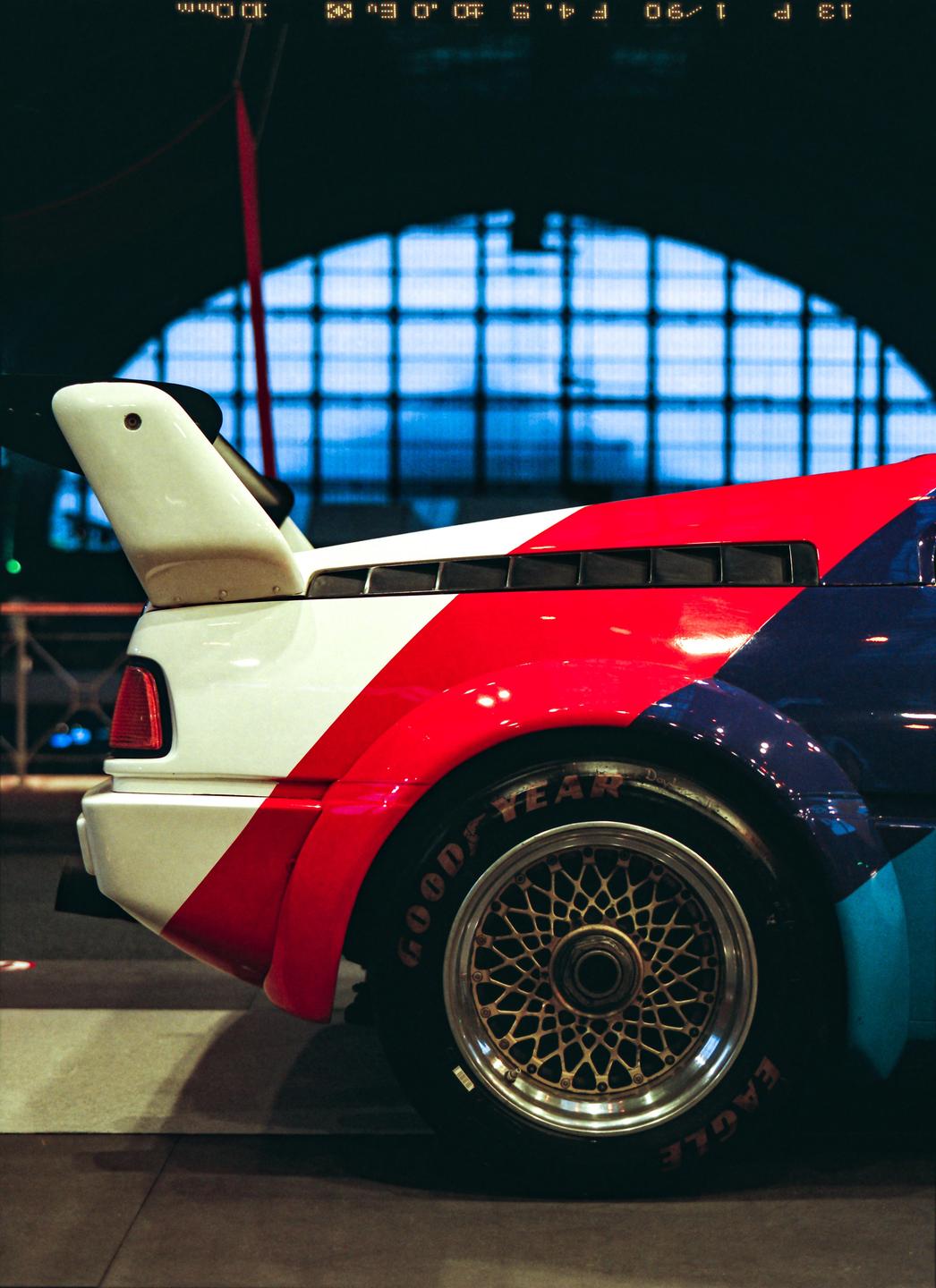 The rear of a red, white, and blue race car with a large spoiler is visible, with a large arched window in the background.