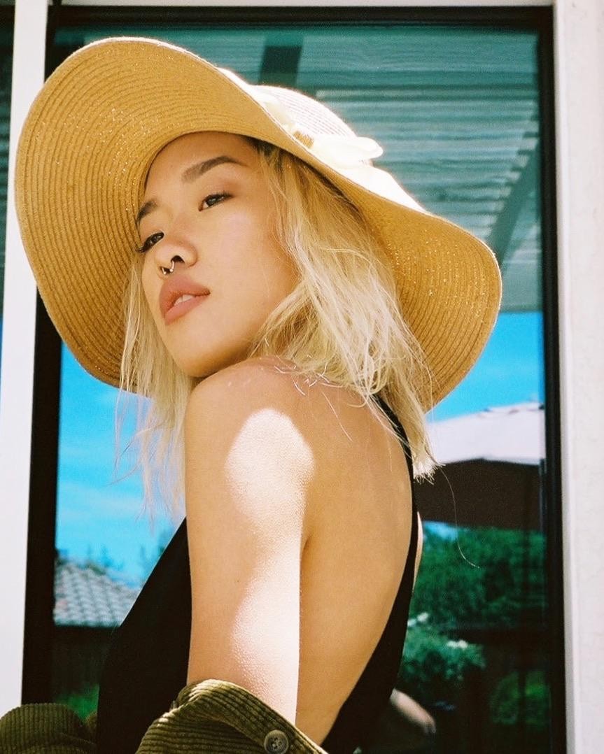 A woman wearing a wide-brimmed hat and a black top looks over her shoulder with a blue sky and green foliage reflected in a window behind her.