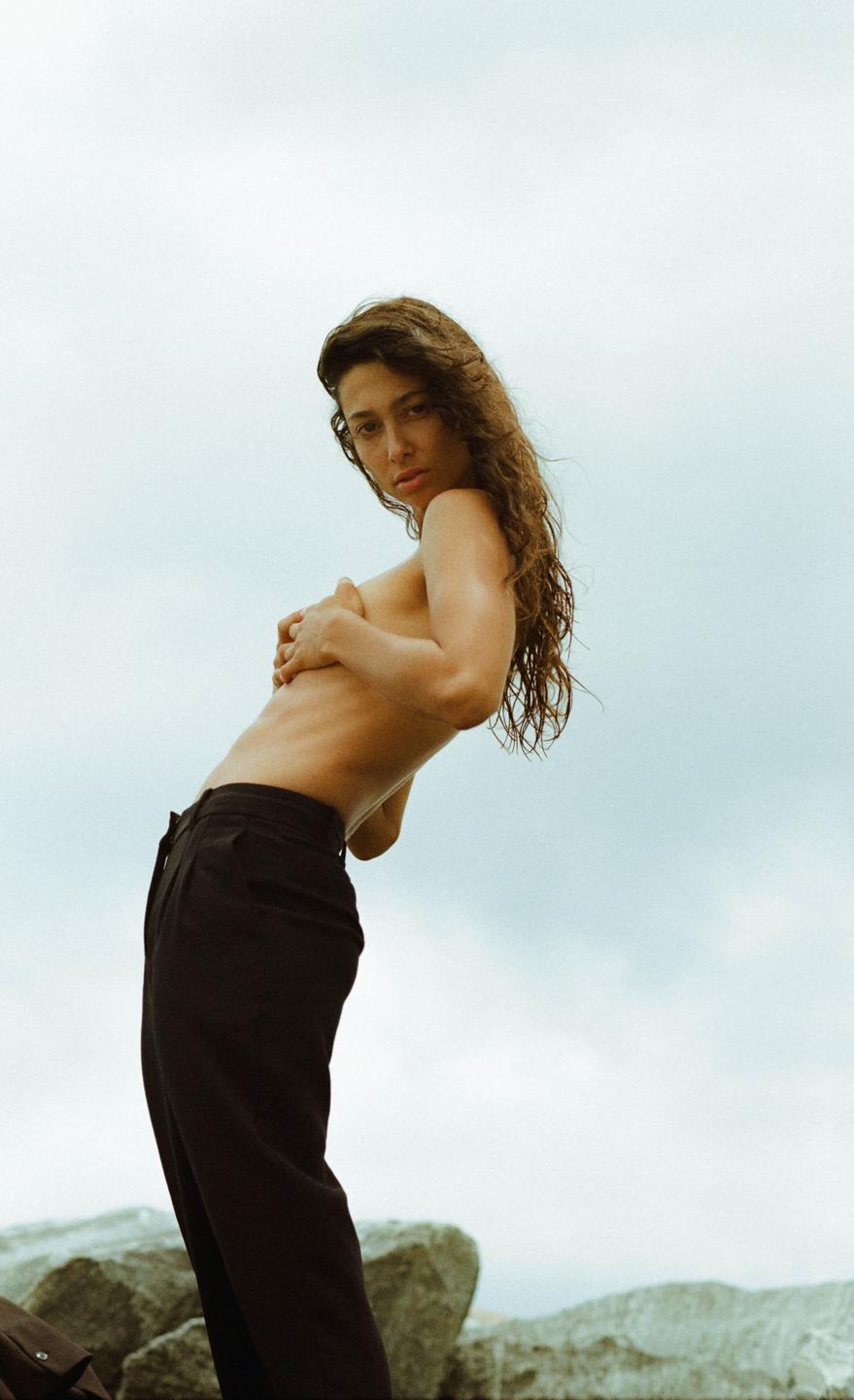 A woman with long, curly hair poses topless in black pants against a light blue sky.