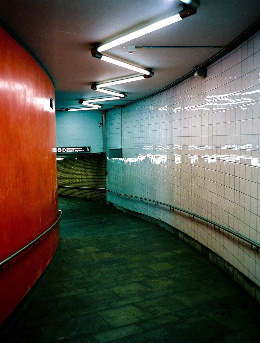 A curved underground walkway is lit by zig-zagging fluorescent lights on the ceiling.