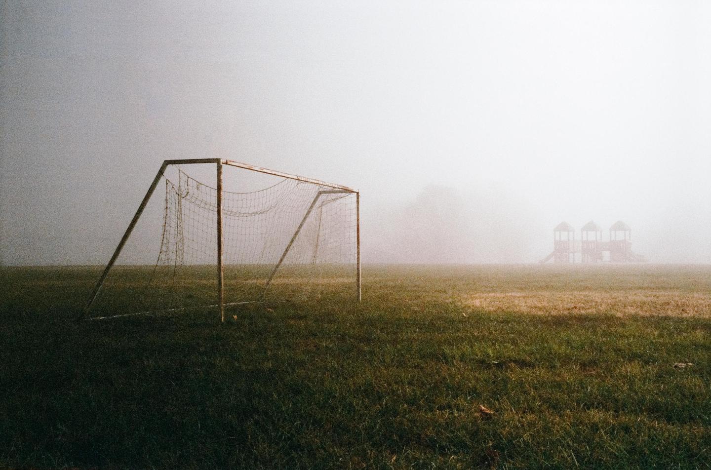 A soccer goal stands on a grassy field with a playground visible in the distance through a thick fog.
